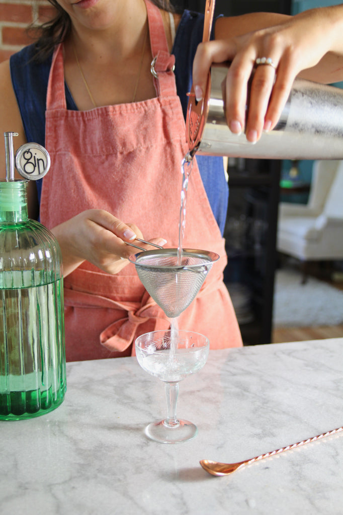 a woman pouring a martini from a cocktail tin into a stemmed glass